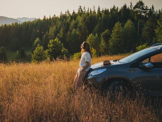Eine Frau, die ihr Auto in einer Berglandschaft geparkt hat und sich an die Motorhaube anlehnt